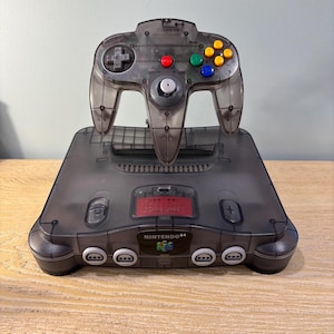 Könnte beinhalten: Eine transparente, graue Nintendo 64 Konsole und ein Controller. Die Konsole trägt den Schriftzug "NINTENDO 64" und ein rotes Kontrolllicht. Der Controller hat farbige Knöpfe und einen Joystick.