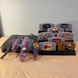 May include: A clear purple Nintendo 64 controller with colorful buttons sits next to a black console and a black game cartridge holder. The holder contains several game cartridges, including "Zelda" and "Mario".