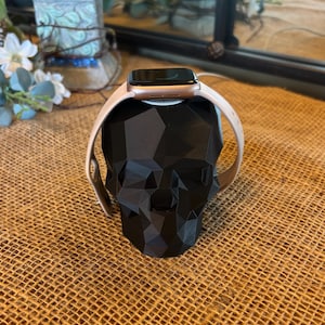 May include: A black geometric skull-shaped watch stand holding a smartwatch with a light pink band. The stand is made of a matte black material and sits on a textured, tan surface. The watch face is black.