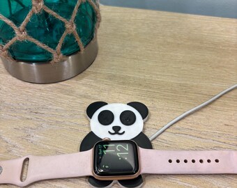 May include: A pink Apple Watch with a black screen displaying the time, held by a panda-shaped charging stand. The stand is black and white. A teal glass jar with a rope net is in the background.