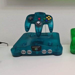 May include: A transparent teal Nintendo 64 console and controller. The controller has a directional pad, colored buttons, and a joystick. The console has the Nintendo 64 logo and controller ports. The set is displayed on a white surface.