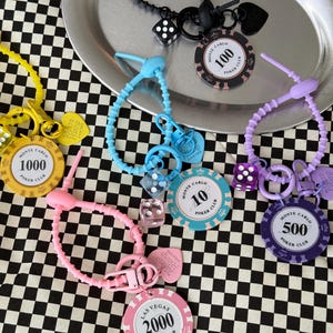 May include: A selection of poker chip keyrings in various colours, including yellow, pink, blue, purple, and black. Each keyring features a poker chip with different denominations and the words "Monte Carlo Poker Club" or "Las Vegas Poker Club."