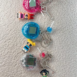 Könnte beinhalten: Drei farbenfrohe Schlüsselanhänger mit Retro-Digital-Haustierspielgeräten in Pink, Blau und Transparent. Jeder Schlüsselanhänger enthält eine kleine Cartoonfigur und einen silberfarbenen Schlüssel- oder Herzanhänger. Der Hintergrund ist eine strukturierte, cremefarbene Oberfläche.