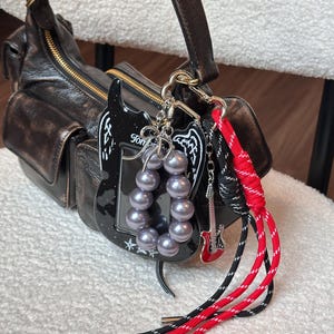 以下が含まれることがあります： ゴールドのジッパーと複数のポケットが付いた黒い革のハンドバッグ。白い翼が付いた黒いギター型のチャーム、グレーのパール、赤と黒のコードなど、いくつかのチャームが取り付けられています。