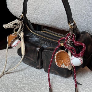 May include: A distressed brown leather shoulder bag with multiple pockets and gold zippers. The bag is adorned with baseball-themed charms, including a baseball, a glove, and a bat, attached with cream and burgundy cords.