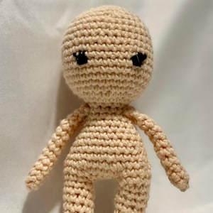 Könnte beinhalten: Eine beige gehäkelte Puppe mit rundem Kopf, schwarzen Augen und einfachen Gliedmaßen. Der Körper der Puppe ist einfarbig, und die Stiche sind deutlich sichtbar. Die Puppe steht aufrecht, mit ausgestreckten Armen.