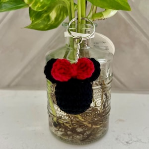 May include: A black crochet keychain shaped like a cartoon mouse head with a red bow. The keychain is attached to a silver metal ring. The background includes a glass vase with a plant inside.