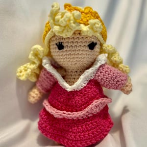 Könnte beinhalten: Handgefertigte Häkelpuppe einer Prinzessin mit einem rosa Kleid und einem weißen Kragen. Die Puppe hat blonde Locken und eine gelbe Krone. Das Gesicht der Puppe hat schwarze Augen und einen hellen Hautton.
