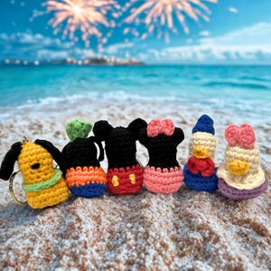 Könnte beinhalten: Eine Sammlung farbenfroher, gehäkelter Charakter-Schlüsselanhänger, darunter ein gelber Hund, Mickey und Minnie Mouse sowie Donald und Daisy Duck. Die Schlüsselanhänger werden an einem Sandstrand mit blauem Meer und Feuerwerk im Hintergrund präsentiert.
