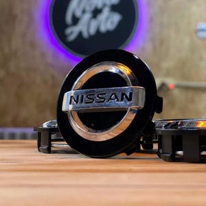 May include: Black and silver Nissan car wheel center cap. The cap has a black background with a silver Nissan logo in the center. The cap is round with a silver ring around the edge.