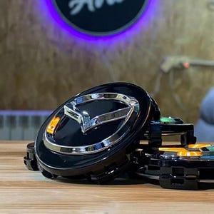 May include: Black and chrome car emblem featuring the Mazda logo. The emblem is circular with a glossy finish and a silver border. The background is blurred, with a wooden surface in the foreground.