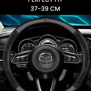 May include: Black steering wheel cover with a carbon fibre texture, designed for a perfect fit. The cover is for wheels sized 37-39 cm. The Mazda logo is visible. The text "STEERING WHEEL COVER PERFECT FIT" is at the top.