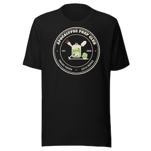 以下が含まれることがあります： 胸元に円形のグラフィックが施された黒の半袖Tシャツ。デザインには「APOCALYPSE PREP CLUB」、「EST. 2026」、「SNACKS PACKED」、「EXITS MAPPED」という文字が含まれており、バックパック、シャベル、手のイラストが描かれています。