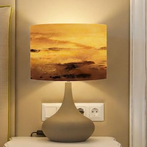 May include: A bedside lamp with a lampshade featuring a landscape painting in shades of yellow, brown, and orange. The lamp has a gray, teardrop-shaped base and sits on a white nightstand. The background includes a bed and a wall.