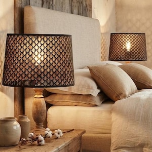 May include: A table lamp with a dark brown, diamond-patterned shade and a turned wooden base. The lamp sits on a rectangular wooden side table next to a bed with beige linen bedding, a knit throw blanket, and a tall, light-coloured headboard.