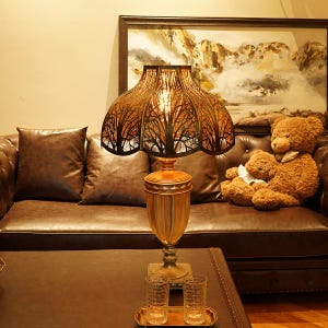 May include: A decorative table lamp with a bronze-colored base and a lampshade featuring a tree branch design. The lamp is on a brown table, with two glasses on a tray in front of it. A brown leather couch and a teddy bear are in the background.