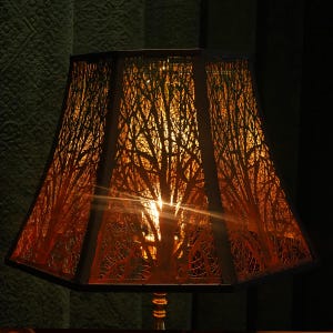 May include: A lampshade featuring a tree branch pattern. The lampshade has a warm orange hue, set within a dark brown frame. Light emanates through the detailed cut-out design, creating a warm atmosphere. This is a home decor item.
