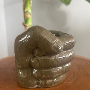May include: A ceramic fist-shaped planter with a speckled, earthy brown glaze. This unique decorative item is ideal for small plants or succulents. The clenched fist shows the knuckles and details of the hand clearly.