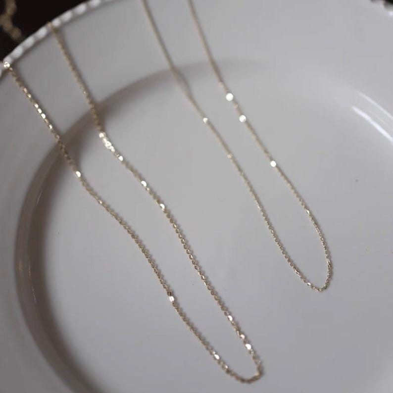 Peut inclure: Deux colliers en or d&eacute;licats avec de fines cha&icirc;nes sont pr&eacute;sent&eacute;s sur une assiette blanche. Les colliers ont un &eacute;clat subtil, captant la lumi&egrave;re. Les cha&icirc;nes sont fines et &eacute;l&eacute;gantes, sugg&eacute;rant un style de bijoux minimaliste.
