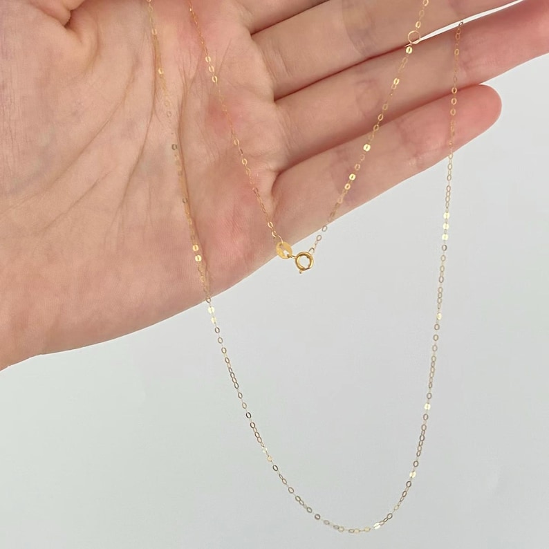Peut inclure: D&eacute;licat collier en or avec un petit fermoir. La cha&icirc;ne est compos&eacute;e de petits maillons rectangulaires plats. Le collier est tenu dans une main sur fond blanc. Le collier est un bijou simple et &eacute;l&eacute;gant.