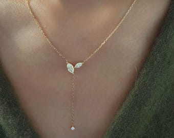 Puede incluir: Un delicado collar de oro con tres piedras preciosas claras y brillantes. Dos piedras con corte marquesa están colocadas una al lado de la otra, con una sola piedra cuadrada colgando debajo. El collar se muestra sobre un fondo verde.