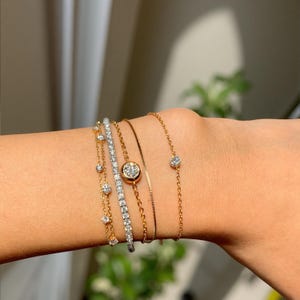 May include: Several gold and silver bracelets, each adorned with sparkling diamond details. The bracelets feature different designs, including fine chains with individual stones and a tennis bracelet style. The jewellery is shown on a wrist.