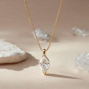 Könnte beinhalten: Eine goldene Halskette mit einem Anhänger aus einem Diamanten im Marquiseschliff. Der Diamant ist in einer goldenen Fassung gefasst und hängt an einer feinen Goldkette. Der Hintergrund ist ein weiches Beige mit dekorativen Steinen.