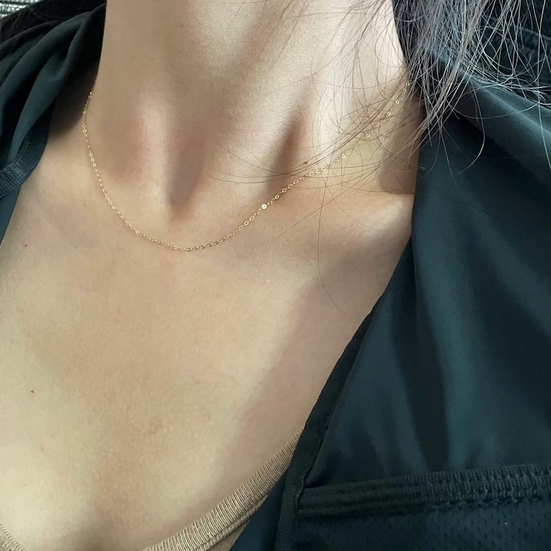 Peut inclure: Un d&eacute;licat collier dor&eacute; reposant sur la poitrine d'une personne. Le collier est une cha&icirc;ne simple, avec de petits maillons espac&eacute;s r&eacute;guli&egrave;rement. L'arri&egrave;re-plan comprend une veste vert fonc&eacute; et un haut beige.