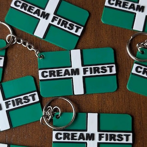 Pode incluir: Chaveiros verdes e brancos com a inscrição "CREAM FIRST". Os chaveiros têm formato retangular com uma cruz branca e o texto em branco. Os chaveiros são presos a argolas prateadas.