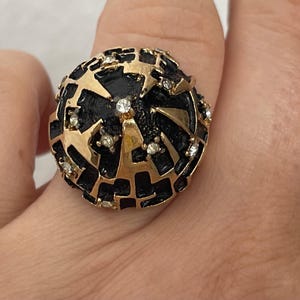 May include: A gold-tone ring featuring a large, rounded, dome-shaped top with a textured black enamel finish. The surface is overlaid with a raised, abstract gold-tone geometric lattice pattern and accented with small, clear, round-cut rhinestones.