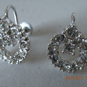 May include: A pair of silver-toned screw-back earrings. Each earring features a circular design encrusted with numerous clear, sparkling rhinestones. The earrings have a vintage aesthetic.