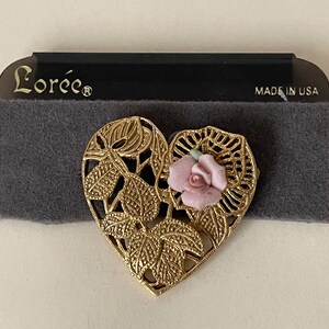 May include: A gold heart-shaped brooch with a pink porcelain rose and intricate leaf details. The brooch is on a gray velvet background and has the text "Lorée" and "Made in USA" on a black label.