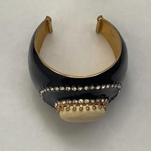 Puede incluir: Un brazalete ancho con un acabado de esmalte negro brillante sobre una base de metal dorado. El centro est&aacute; adornado con una piedra ovalada de color crema rodeada por un borde de brillantes transparentes engastados en garras de metal dorado.