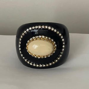 Puede incluir: Una pulsera brazalete ancha de color negro con una piedra cabuj&oacute;n ovalada de color crema en el centro. La piedra est&aacute; rodeada por un borde de metal dorado y un anillo exterior de peque&ntilde;os brillantes claros sobre la superficie negra lisa.