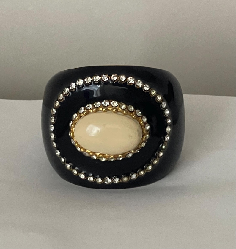 Puede incluir: Una pulsera brazalete ancha de color negro con una piedra cabuj&oacute;n ovalada de color crema en el centro. La piedra est&aacute; rodeada por un borde de metal dorado y un anillo exterior de peque&ntilde;os brillantes claros sobre la superficie negra lisa.