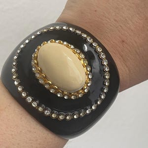 Puede incluir: Una pulsera brazalete ancha de color negro con un cabuj&oacute;n central ovalado de color crema engastado en un marco dorado. La pulsera est&aacute; decorada con dos c&iacute;rculos conc&eacute;ntricos de brillantes transparentes incrustados en la superficie negra lisa.