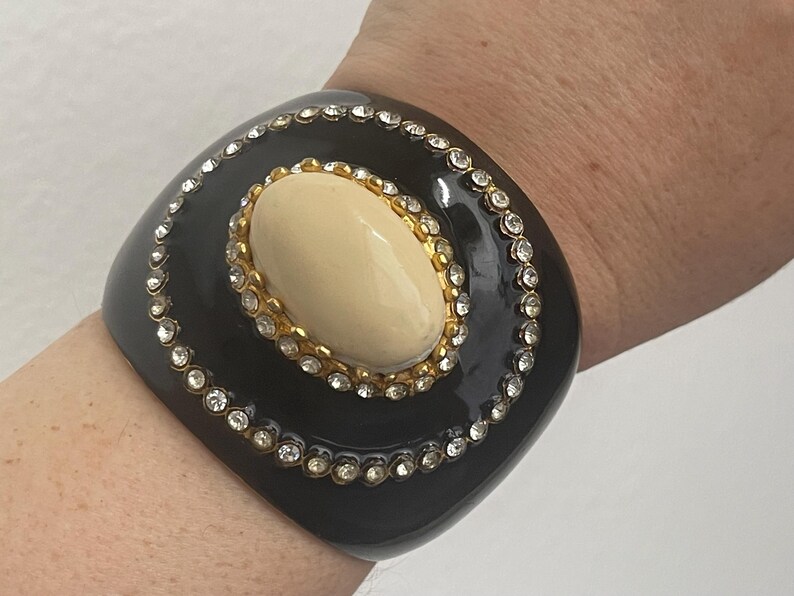 Puede incluir: Una pulsera brazalete ancha de color negro con un cabuj&oacute;n central ovalado de color crema engastado en un marco dorado. La pulsera est&aacute; decorada con dos c&iacute;rculos conc&eacute;ntricos de brillantes transparentes incrustados en la superficie negra lisa.