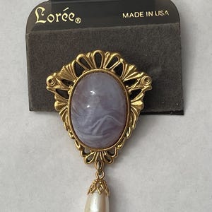 May include: A gold-toned brooch with an oval, marbled, lavender-colored center stone. The brooch has a decorative filigree frame and a dangling pear-shaped faux pearl. The backing card reads "Lorée" and "MADE IN USA."