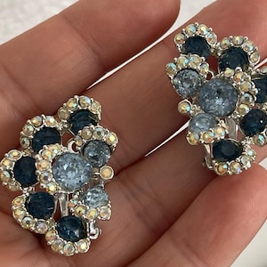 May include: A pair of vintage clip-on earrings. Each earring features a floral design with a cluster of blue and iridescent crystals. The earrings have a silver-tone metal setting.