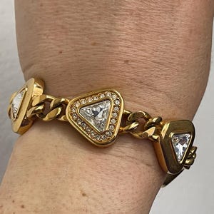 May include: Gold-toned bracelet featuring triangular and square-shaped links. Each link is adorned with a large, clear, triangular crystal surrounded by smaller, clear crystals. The bracelet has a chain-link design.