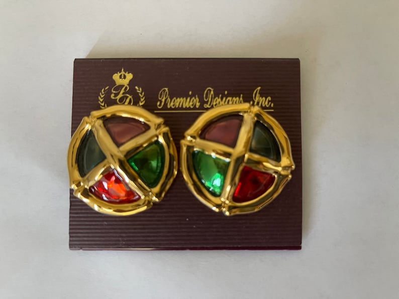 May include: A pair of gold tone earrings with a round, multi-colored glass design. The earrings are on a brown card with the text "Premier Designs Inc."