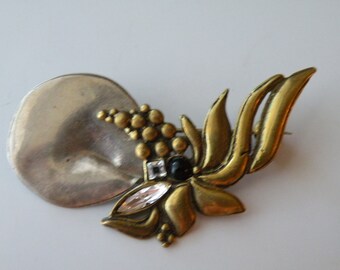 May include: A vintage brooch with a silver-toned, circular base and a gold-toned floral design. The brooch features a cluster of small, gold-toned spheres, a black gem, and a clear, elongated gem. The pin is a unique accessory.