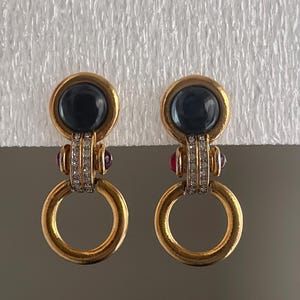 May include: A pair of gold-toned drop earrings featuring a large, smooth, dark blue cabochon stone at the top, accented by a vertical row of clear crystals, small red side stones, and a circular gold ring dangling at the bottom of each piece.