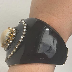 Puede incluir: Una pulsera brazalete ancha de color negro brillante con una piedra cabuj&oacute;n central elevada de color crema, montada en un engaste dorado en forma de corona. La piedra est&aacute; rodeada por una l&iacute;nea curva de brillantes transparentes incrustados.
