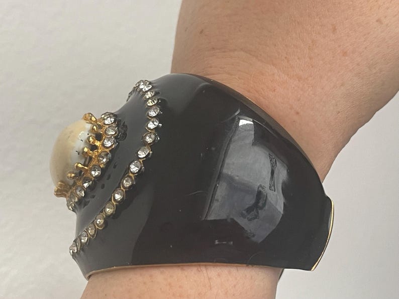 Puede incluir: Una pulsera brazalete ancha de color negro brillante con una piedra cabuj&oacute;n central elevada de color crema, montada en un engaste dorado en forma de corona. La piedra est&aacute; rodeada por una l&iacute;nea curva de brillantes transparentes incrustados.