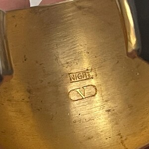 Puede incluir: Un brazalete ancho con un interior de metal dorado pulido y paneles exteriores negros brillantes. La superficie interior est&aacute; grabada con el texto NIGHT y un logotipo V dentro de un contorno ovalado, mostrando un acabado met&aacute;lico y textura lisa.