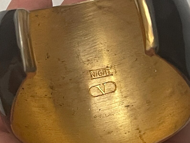 Puede incluir: Un brazalete ancho con un interior de metal dorado pulido y paneles exteriores negros brillantes. La superficie interior est&aacute; grabada con el texto NIGHT y un logotipo V dentro de un contorno ovalado, mostrando un acabado met&aacute;lico y textura lisa.