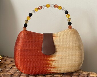 May include: A woven handbag with a brown leather flap and a handle made of colorful beads. The bag is a gradient of orange and beige.