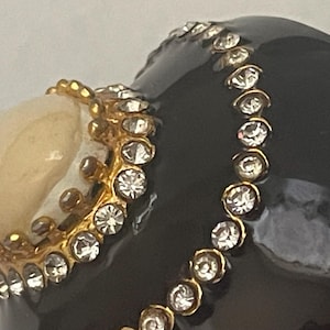Puede incluir: Un objeto decorativo negro brillante con una superficie lisa adornada con un borde de metal dorado incrustado con pedrer&iacute;a transparente. Una piedra cabuj&oacute;n redondeada de color blanco hueso est&aacute; engastada en una base de metal dorado en forma de corona.