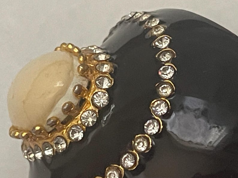Puede incluir: Un objeto decorativo negro brillante con una superficie lisa adornada con un borde de metal dorado incrustado con pedrer&iacute;a transparente. Una piedra cabuj&oacute;n redondeada de color blanco hueso est&aacute; engastada en una base de metal dorado en forma de corona.